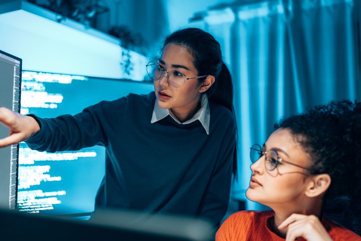 Two women focused on a computer screen in a blue-lit room.