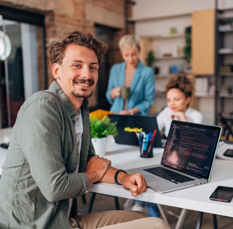 A smiling man working on a laptop in a cozy office space.