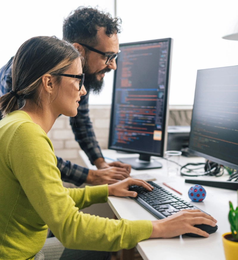 Two developers collaborating on coding at dual monitors in an office.